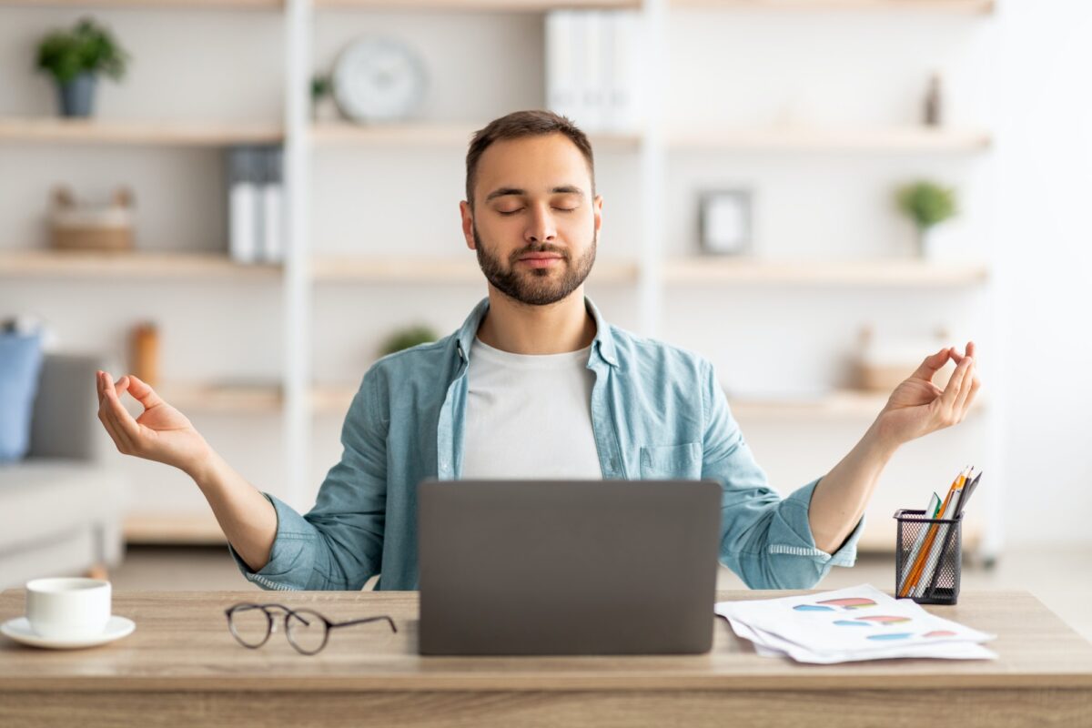 Workplace stress management. Calm Caucasian man meditating with closed eyes in front of laptop pc at